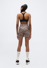 Γυναίκα που στέκεται με την πλάτη γυρισμένη, φορώντας μαύρο αθλητικό σουτιέν, σορτς με λεοπάρ σχέδιο, λευκές κάλτσες και μαύρα αθλητικά παπούτσια πάνω σε απλό φόντο.