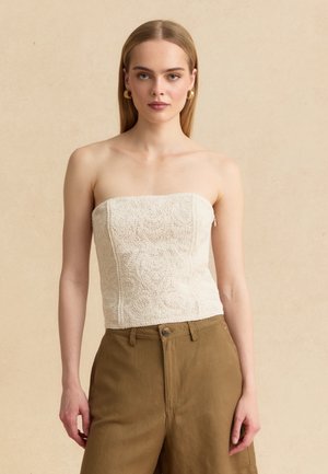 Giovane donna con capelli biondi lisci indossa un top panna in pizzo senza spalline e pantaloni marroni a vita alta, posando su uno sfondo beige.