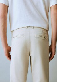 Une personne se tient de dos, portant une chemise blanche et un pantalon beige clair, les mains le long du corps.