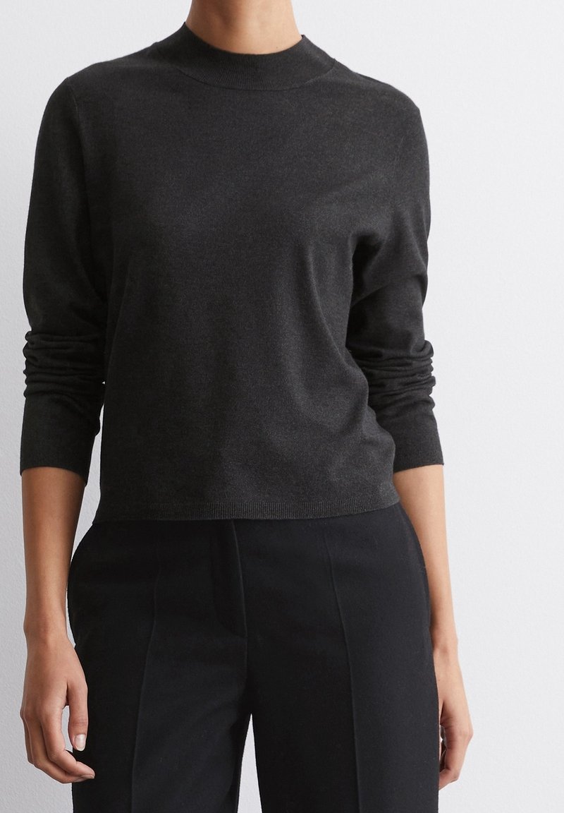 Personne portant un haut ajusté à manches longues gris foncé avec un col montant et un pantalon noir ajusté sur un fond blanc uni.