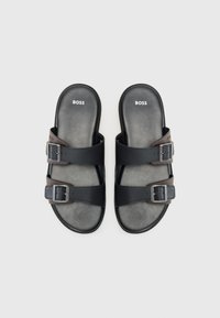 Sandales noires en cuir avec deux sangles réglables à boucles. Semelle en caoutchouc grise, texture lisse et logo de la marque en relief en blanc sur le semelle.