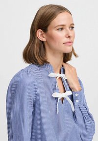 Chemise à rayures bleues et blanches à manches longues avec un col montant, présentant des liens en ruban blanc et des poignets à boutons. Tissu en coton lisse.
