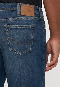 Dark blue denim jeans with a straight cut, featuring a leather patch at the waistband and a curved stitching pattern on the back pocket.