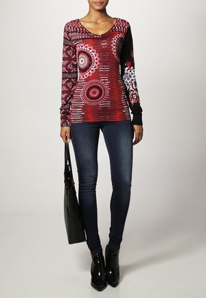 Kobieta ubrana w czerwoną, wzorzystą bluzkę z długim rękawem, ciemne obcisłe jeansy, czarne botki na kostkę, trzymająca czarną torebkę na tle neutralnym.