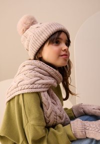 Knit beige beanie with pom-pom, matching scarf with cable pattern, green sweater, and beige fingerless gloves, seated against a neutral background.