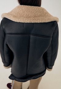 Veste en cuir noir avec un col en peau retournée beige, présentant une forme structurée et des coutures apparentes à l'arrière.