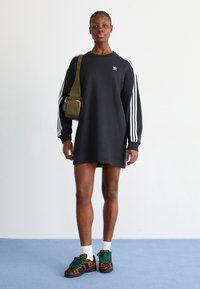 Svart sweatshirtklänning med vita ränder på ärmarna, i kombination med en grön axelväska och mönstrade sneakers. Strukturerad tyg, avslappnad passform.