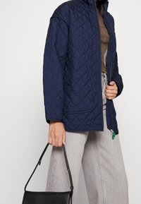 Chaqueta acolchada navy con cremallera completa, dos bolsillos frontales y un acolchado ligero, combinada con pantalones grises y un bolso negro.