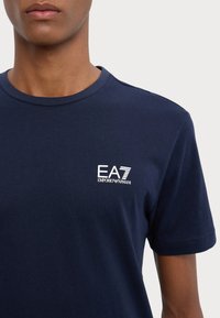 Tamsiai mėlynas medvilninis marškinėliai su apvaliu kaklu, trumpomis rankovėmis ir baltu EA7 Emporio Armani logotipu kairėje krūtinės pusėje.