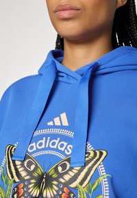 Plava pamučna dukserica ima središnji grafički prikaz leptira u živopisnim bojama, kontrastne vezice i Adidas logo na prsima.