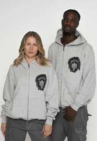 Graue Zip-Hoodies mit Fronttaschen, großen Grafikakzenten eines Löwenkopfes und dem Text "MULTIPLY". Weicher Stoff mit lockerer Passform.