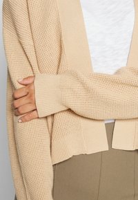 Beżowy sweter właśnie z dzianiny, z teksturowaną powierzchnią i ściągaczami u mankietów. Otwarty przód, odsłaniający warstwowy wygląd na białej bluzce.