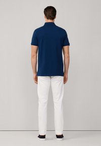 Polo bleu marine à manches courtes, porté avec un pantalon blanc slim et des chaussures décontractées navy, vu de dos sur un fond neutre.