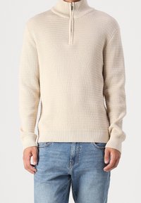 Pull en tricot beige avec un col haut et une demi-fermeture éclair. Tissu texturé, poignets et ourlet côtelés, associé à un jean bleu.