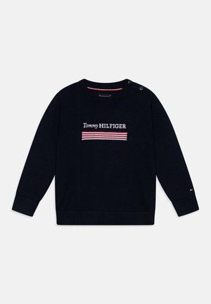 Granatowy, długi rękaw, dziany sweter z napisem „Tommy Hilfiger” i czerwono-białym paskowanym wzorem na piersi, z zapięciem na guziki na ramieniu.