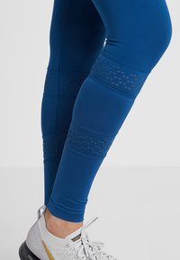 Blå leggings i stretchmaterial har nätutskärningar med geometriska mönster vid vadarna. Kombineras med vita träningsskor.