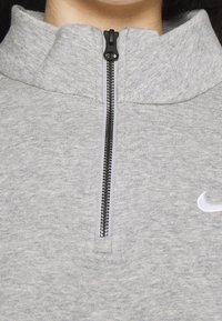 Grå zip-hoodie med hög krage, svart dragkedja och en liten vit Nike-logotyp på vänster bröst. Mjuk, texturerad tyg.