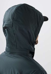 Homme portant une veste à capuche imperméable foncée avec une fermeture éclair ajustable et une casquette noire sous la capuche, de profil.
