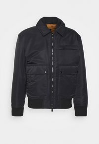 Svart bomberjacka i slät tyg. Har dragkedja framtill, ribbade muddar, två framfickor och en bröstficka. Insidan är fodrad i brunt.