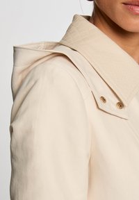 Veste beige avec un col texturé de couleur claire. Elle présente des accents en or et une texture de tissu lisse. Coupe ajustée avec un design structuré.