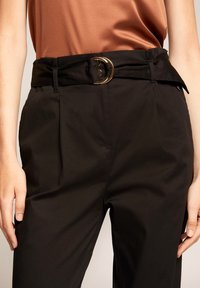 Svarta skräddarsydda byxor har hög midja med veck och ett brett bälte. Bältet har ett guld-färgat, runt spänne som accent. Materialet har en slät textur.