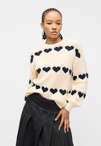 Γυναίκα με πλεξούδες που φοράει εκρού πουλόβερ με μοτίβα μαύρης καρδιάς και πλισέ μαύρη φούστα από denim, ποζάροντας μπροστά σε λευκό φόντο.