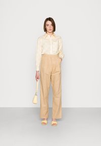 Camisa abotoada em amarelo claro com um padrão quadriculado, combinada com calças de cintura alta em bege. Acessórios incluem uma pequena bolsa amarela e chinelos.