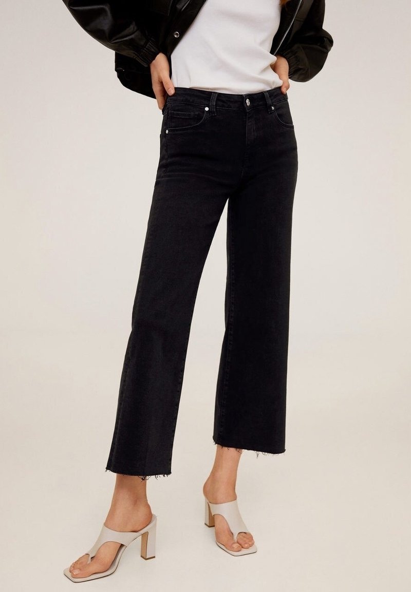 Schwarze Jeans mit weitem Bein aus Denim und ausgefranstem Saum, kombiniert mit hellen Sandalen mit blockförmigem Absatz. Einfaches weißes Oberteil und schwarze Jacke.