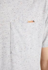 Světle šedé tričko s tečkovaným vzorem a malou kapsou, na které je vyšitý design osoby opalující se vedle žraločí ploutve.