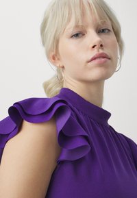 Top mov cu mâneci volănașe, având un guler înalt și un design plisat. Țesătură fină cu o textură moale și un luciu subtil.