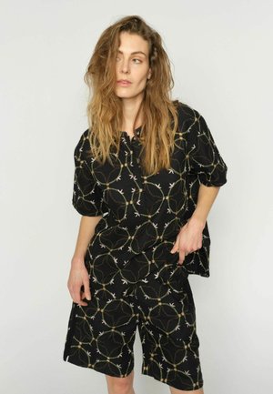 Femeie cu păr lung și ondulat, purtând un costum negru format din cămașă și pantaloni scurți asortați, cu model circular și de ramuri, stând în fața unui fundal simplu și deschis la culoare.