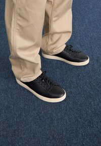 Black leather sneakers with round toe, white rubber sole, flat laces, paired with beige pants on a blue carpeted surface.