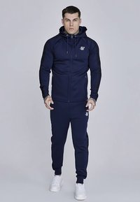 Granatowy dres z bluzą na zamek i zwężanymi spodniami. Charakterystyka: logo, dopasowany krój i ściągacze na mankietach. Tkanina wydaje się gładka i trwała.