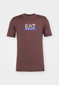 Brun kortermet T-skjorte laget av glatt stoff, med en hvit og blå "EA7 Emporio Armani" logo foran. sømdetaljer synlige.