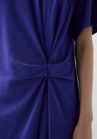 Paarse jurk met een gedrapeerd ontwerp en een gedraaide knoopdetail in de taille, gemaakt van zachte stof met een gladde textuur en een gematigde glans.