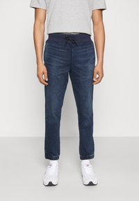 Man som bär mörkblå joggerjeans med elastisk midja och mudd, i kombination med vita sneakers och grå t-shirt.