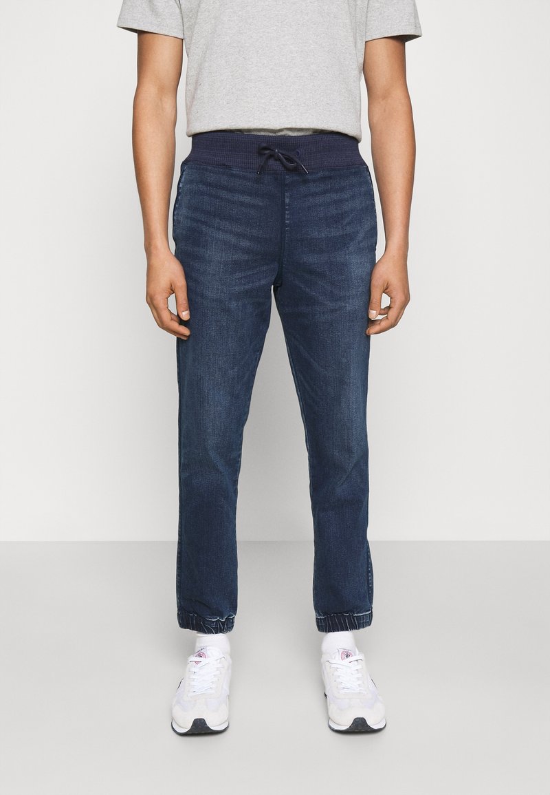 Man som bär mörkblå joggerjeans med elastisk midja och mudd, i kombination med vita sneakers och grå t-shirt.