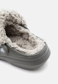 Chausson gris avec une doublure douce et duveteuse beige. Forme arrondie, semelle extérieure texturée et accent de logo circulaire sur le côté.