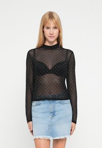 Sort netbluse med lange ærmer og høj hals, pyntet med sølvfarvede polkaprikker, lagdelt over en sort bh, kombineret med en lys denim mininederdel.