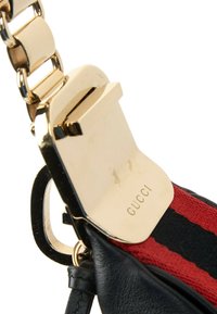 Gros plan d'un fermoir doré gravé Gucci sur cuir noir et une sangle en tissu rayé rouge et noir avec un maillon de chaîne doré.