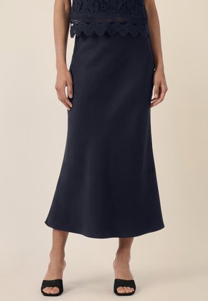 Frau trägt einen marineblauen Rock bis zum Knöchel, kombiniert mit einem schwarzen, offenen Sandalettenabsatz, stehend vor einem schlichten Hintergrund.