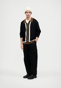 Fekete gombos kardigán fehér és barna részletekkel, textúrált kötött anyagból. Fekete cargo nadrággal és fekete cipővel párosítva, egy krémszínű sapkával a tetején.