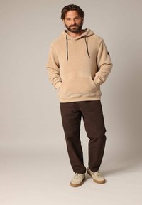 Beige fleece hoodie met een kangoeroe-pocket en zwarte trekkoorden, gecombineerd met donkerbruine broek en lichte schoenen. Effen achtergrond.