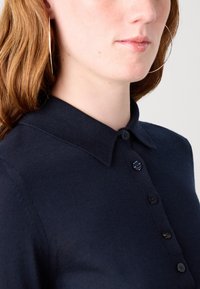 Polo din tricot de culoare bleumarin, cu guler, având o un placket cu șase nasturi negri și detalii vizibile de cusătură pe umăr.