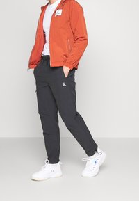 Oranje zip-up jack met een wit logo-embleem, gecombineerd met zwarte broek met zijzakken en een wit logo. Witte sneakers maken de look compleet.