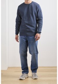 Sudadera azul marino con cuello redondo y puños acanalados, combinada con jeans azul claro y zapatillas de running grises sobre un suelo de madera.