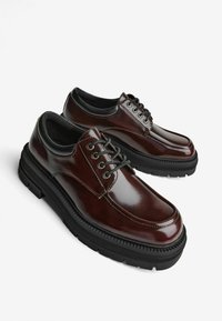 Scarpe in pelle lucida marrone con punte tonde, suole in gomma nera e lacci neri. Presentano una texture liscia e dettagli di cucitura lungo i lati.