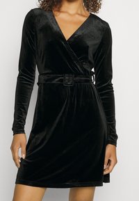 Svart sammetsomslagsklänning med långa ärmar, V-ringning, insvängd midja med bälte, slät textur och längd ovanför knäna.