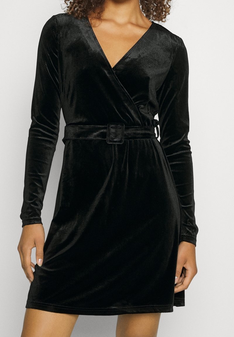 Svart sammetsomslagsklänning med långa ärmar, V-ringning, insvängd midja med bälte, slät textur och längd ovanför knäna.