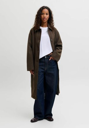 Femme aux cheveux bouclés portant un long manteau vert olive, un t-shirt blanc, un jean large bleu foncé et des chaussures foncées, debout avec une main dans la poche.